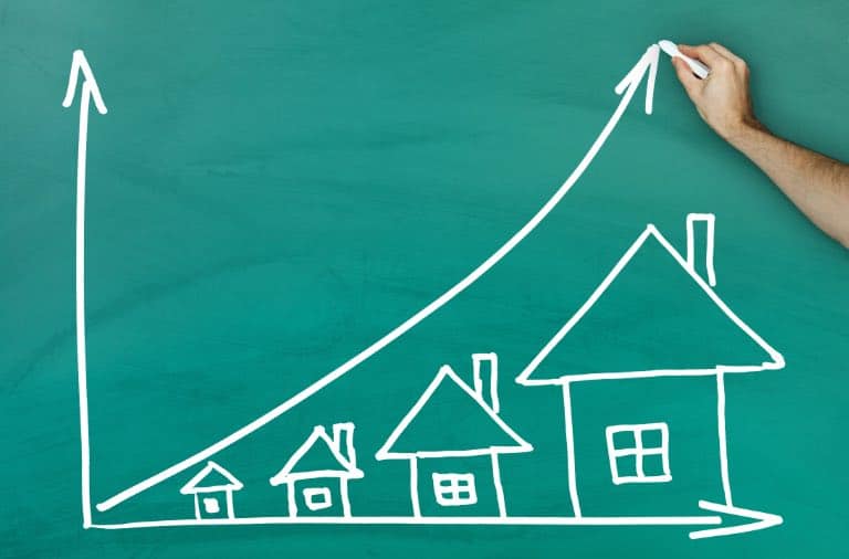 A hand drawing a graph on a blackboard with a house, showcasing the increase in property value to help illustrate why do property prices go up in value.